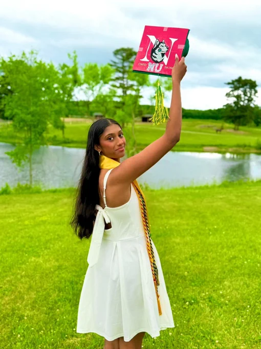 Smiling young woman with long dark hair, in a white dress and yellow/multicolored stole, raises a red graduation cap. The cap reads \"ONTO NU THING\" with a large \"N\" and husky head. She stands in a grassy field with a pond and trees.