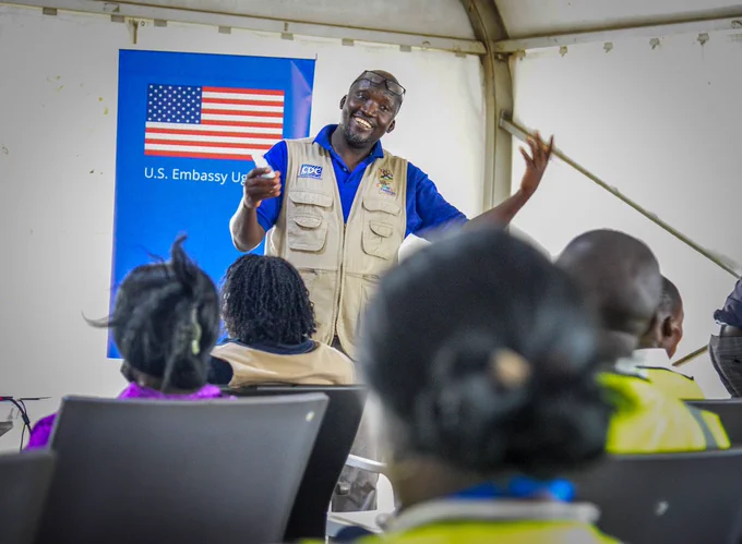 A smiling presenter stands at the front of a training session, speaking and gesturing to an audience seated in rows under a large tent. Behind the speaker is a banner displaying the U.S. flag and the words “U.S. Embassy Uganda.” 