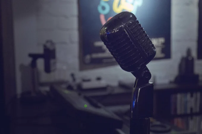 A close-up shot of a vintage-style black microphone on a stand, illuminated by dramatic lighting, set against a blurred background of a music studio with a poster and audio equipment.