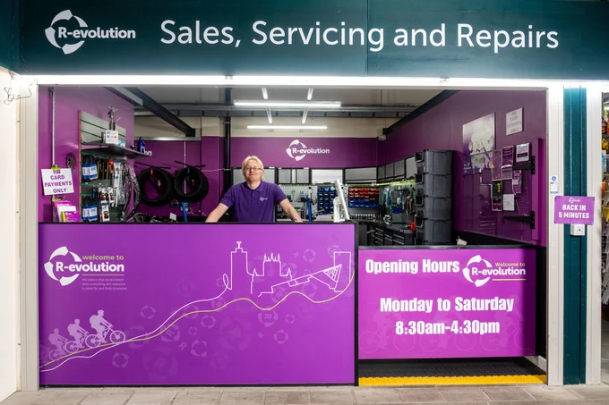 A man stands behind the counter of a purple bicycle shop, 'R-evolution', which sells, services, and repairs bikes.
