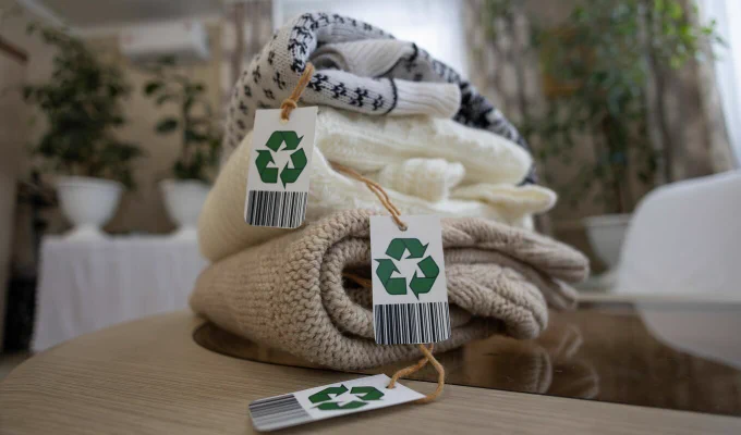 A stack of folded sweaters in various patterns and neutral colors is displayed on a wooden table. Each sweater has a tag featuring a recycling symbol and a barcode. The background includes blurred houseplants and furniture.