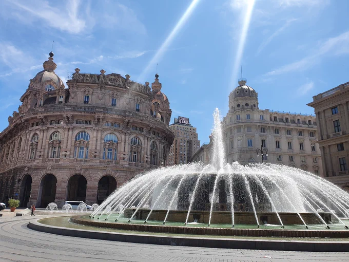 Zu sehen ist ein großer runder Springbrunnen. Zahlreiche kleine Fontänen schießen Wasser nach schräg oben, so dass ein Kreis entsteht. In der Mitte des Kreises ist eine weitere Fontäne, die ihr Wasser gerade nach oben schießt.
Im Hintergrund sind prächtige, reich verzierte Altbauten und ein blauer Himmel zu sehen.