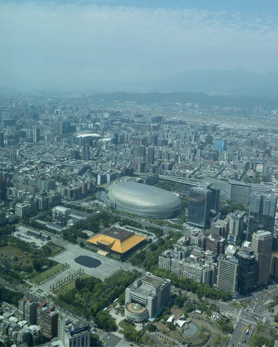 Aerial view of Taipei, featuring the Taipei Dome stadium in the center, surrounded by numerous buildings and roads. Green parks and open spaces are visible throughout the cityscape. The distant mountains frame the skyline under a mostly clear sky.