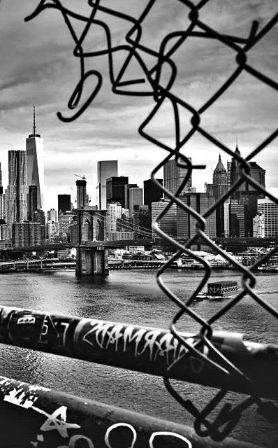 A high-contrast, black and white street photograph of the Lower Manhattan skyline and the Brooklyn Bridge, framed through a torn opening in a chain-link fence. 