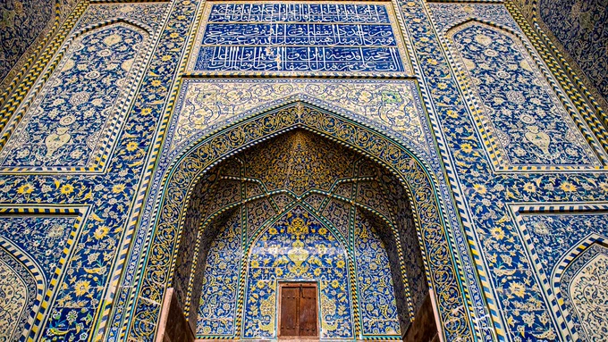 Low-angle view of an intricately tiled, arched entrance inside the Sheikh Lotfollah Mosque in Isfahan, Iran. The structure features magnificent mosaic tilework, dominated by deep cobalt blues, vibrant yellows, and whites, that forms complex floral arabesques and geometric patterns. A large rectangular panel of white Arabic calligraphy on a blue background is positioned directly above the central archway.