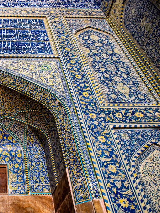 Low-angle view of an intricately tiled, arched entrance inside the Sheikh Lotfollah Mosque in Isfahan, Iran. The structure features magnificent mosaic tilework, dominated by deep cobalt blues, vibrant yellows, and whites, that forms complex floral arabesques and geometric patterns. A large rectangular panel of white Arabic calligraphy on a blue background is positioned directly above the central archway.