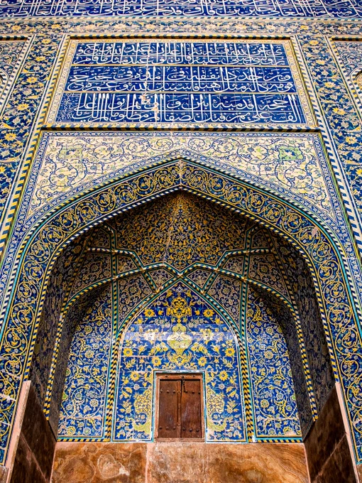 Low-angle view of an intricately tiled, arched entrance inside the Sheikh Lotfollah Mosque in Isfahan, Iran. The structure features magnificent mosaic tilework, dominated by deep cobalt blues, vibrant yellows, and whites, that forms complex floral arabesques and geometric patterns. A large rectangular panel of white Arabic calligraphy on a blue background is positioned directly above the central archway.