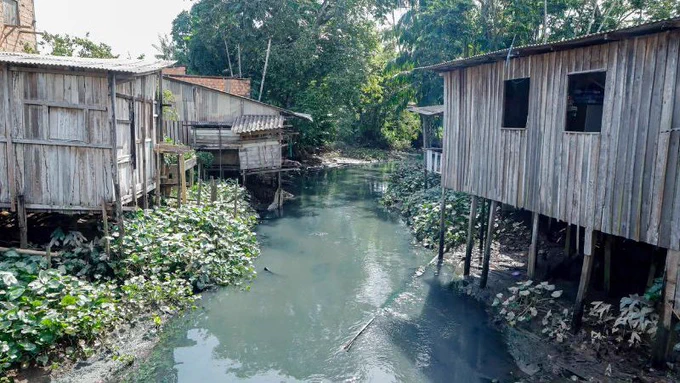 Rio com águas escuras ladeado por casas de madeira suspensas sobre palafitas e vegetação verde ao fundo.