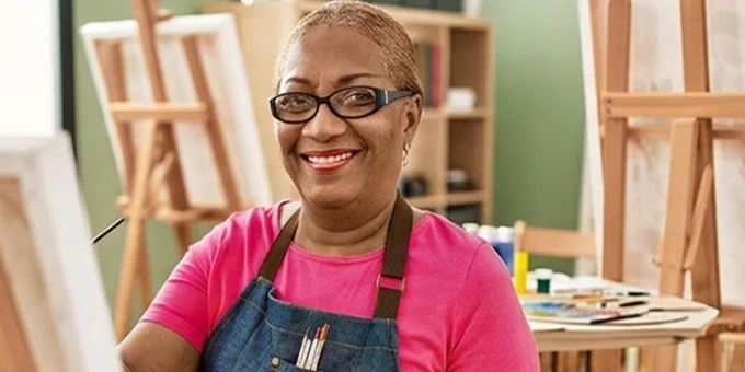 Person wearing a pink shirt and denim apron painting on a canvas in a bright art studio with wooden easels.