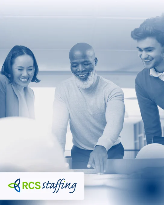 Three diverse colleagues smiling and collaborating together around a desk in a modern office setting, conveying teamwork and positivity.