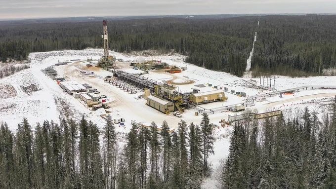Aerial view of a snow-covered oil and gas drilling site surrounded by dense forest. The site features a tall drilling rig, several buildings, and parked vehicles.