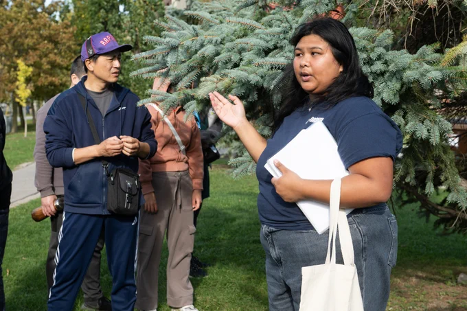 Woman holding papers and a tote bag gestures toward a pine tree while speaking to a group outdoors in a park.