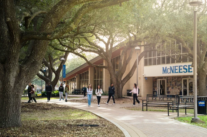 Students walking along a tree-lined pathway in front of the McNeese campus building on a sunny day.