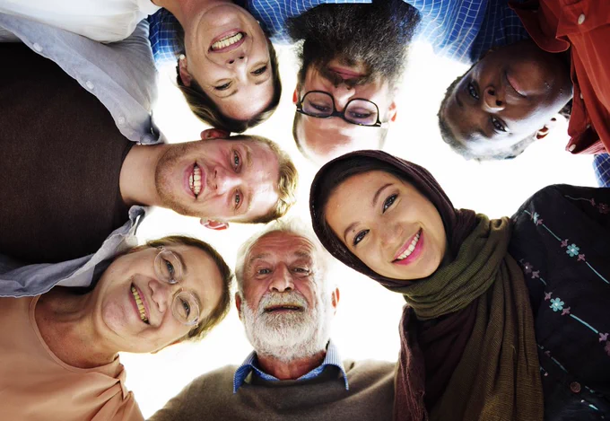 A group of people from a diverse range of ages and backgrounds are standing in a circle, smiling and looking down towards the camera to pose for a photograph.