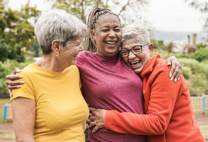 Three older women embracing and laughing joyfully outdoors.