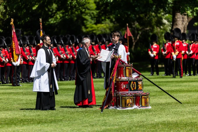 Three chaplains in formal robes stand on the grass grounds of Buckingham Palace as they consecrate the new Colours for the Grenadier Guards at Buckingham Palace. Regimental drums are stacked, draped with regimental flags to form the alter for the drumhead service.  A large formation of guardsmen in red tunics and bearskins are formed behind.