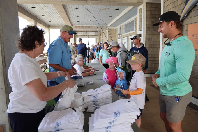A group of people are gathered around a table for a fishing event at Jennette's Pier in Nags Head. On the table are stacked shirts with a fishing club logo. Some participants, including children, are receiving the shirts. Fishing rods are visible in the background.