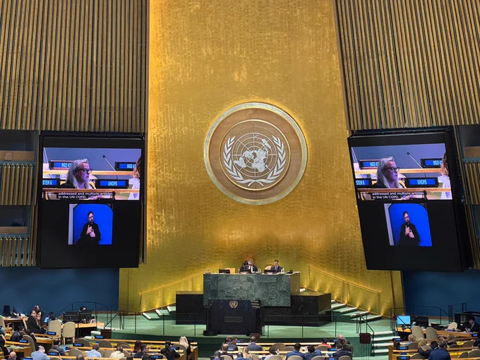 View of the United Nations General Assembly Hall from the delegates’ seating area, with a UNOPS nameplate prominently displayed in the foreground. In the background, delegates are gathered across the chamber beneath the large gold United Nations emblem. Two screens show a speaker addressing the assembly with sign language interpretation. The hall is busy with participants attending the Conference of States Parties to the Convention on the Rights of Persons with Disabilities (COSP19).