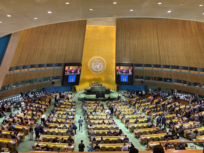 Wide view of the UN General Assembly hall in New York. Rows of curved desks fill the floor, with many delegates seated or moving between aisles. At the front, a raised podium stands beneath a large gold wall bearing the UN emblem. Two large screens show speakers at the podium and a sign language interpreter. Bright ceiling lights illuminate the formal, crowded setting.