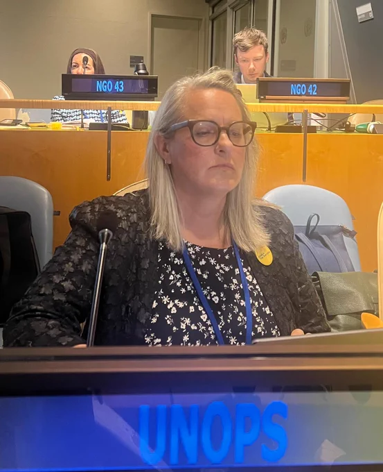 ATscale representative Ceridwen Johnson sits at the UNOPS delegation desk during the 19th Conference of States Parties to the Convention on the Rights of Persons with Disabilities (COSP19) at United Nations Headquarters in New York. She is seated in front of a microphone and laptop, listening attentively during the session. The illuminated UNOPS nameplate is visible in the foreground, with other delegates and NGO representatives seated in the background.