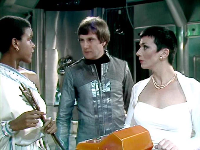 GPT 5.5 describes the image as: "In this scene from the classic BBC Television series Blake’s 7, Josette Simon, Paul Darrow, and Jacqueline Pearce are shown in a sleek, metallic science-fiction interior, suggesting a spacecraft or futuristic control room. Josette Simon stands at left in an elegant white costume with decorative detailing, holding a small fan-like prop. Paul Darrow is centered in a silver-grey, high-collared outfit, turned toward her with a serious, intent expression. Jacqueline Pearce stands at right in a striking white gown with pearls, holding an orange case or container, her gaze directed toward Darrow. The image has the polished, theatrical style of late-1970s/early-1980s BBC sci-fi, with dramatic costumes and a tense, formal atmosphere."
The script search url for this tweet is
https://blake.torpidity.net/s/301/179