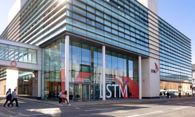 Modern multi-storey glass-fronted building on a city street corner, with large panels of reflective windows and horizontal bands across each floor. The ground level features a tall glass entrance supported by white columns and revolving doors. Prominent signage reads “LSTM” with additional text “Liverpool School of Tropical Medicine” displayed across the frontage. Several pedestrians walk along the pavement in front of the building, and a road with traffic markings runs alongside. A height restriction sign on a nearby walkway reads “4.7m / 15'-6"”. Bright daylight creates reflections on the glass and light shadows on the street.