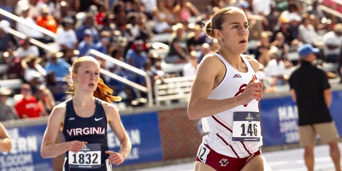 Two athletes are running on a track during a race. The athlete wearing a white and maroon outfit with the number 146 runs ahead, while the athlete in a navy "Virginia" outfit with the number 1832 follows. Spectators are in the background.