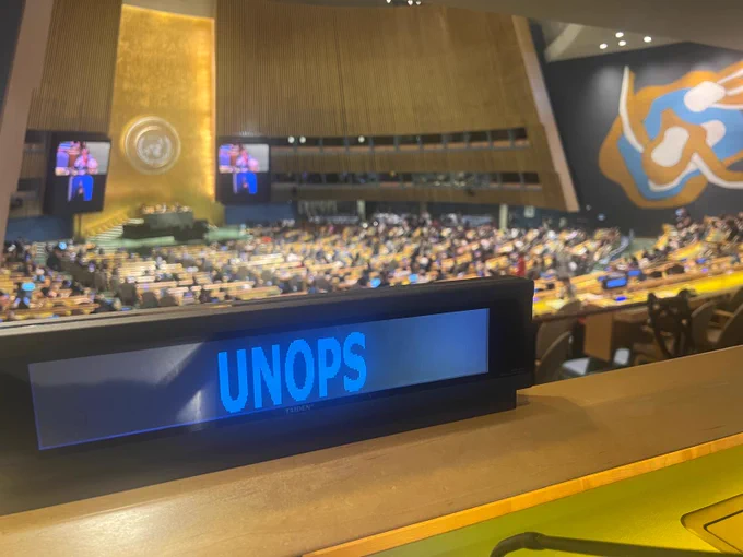 View of the United Nations General Assembly Hall from the delegates’ seating area, with a UNOPS nameplate prominently displayed in the foreground. In the background, delegates are gathered across the chamber beneath the large gold United Nations emblem. Two screens show a speaker addressing the assembly with sign language interpretation. The hall is busy with participants attending the Conference of States Parties to the Convention on the Rights of Persons with Disabilities (COSP19).