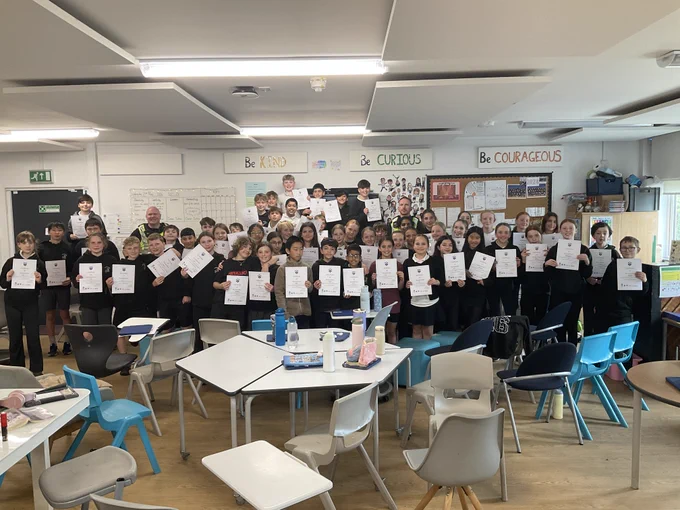A group of schoolchildren and adults proudly display certificates in a classroom decorated with motivational slogans.