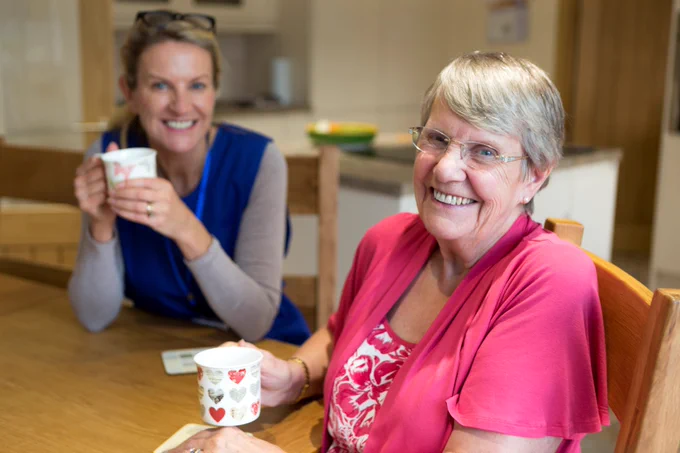 Community care worker joins an an elderly woman for a cup of tea while sitting at her kitchen table