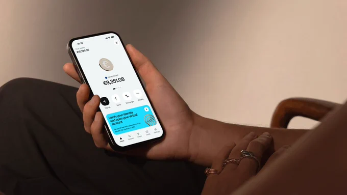 A person holds a smartphone displaying a financial app with a balance of €9,351.08 and options for managing accounts.