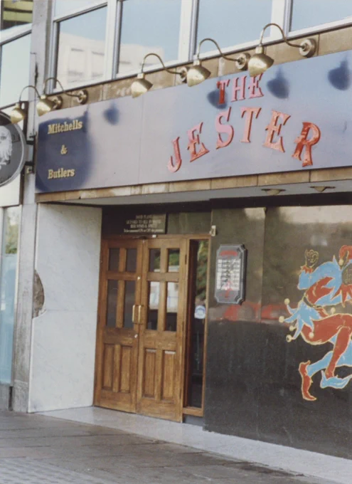 Photograph of the frontage of a public house. Facia above the entrance reads The Jester with drop lighting above. A Mitchells & Butler sign in the top left of the facia. A wooden door on the left beneath the facia with coloured glass on the right with a figure depicting a court jester.