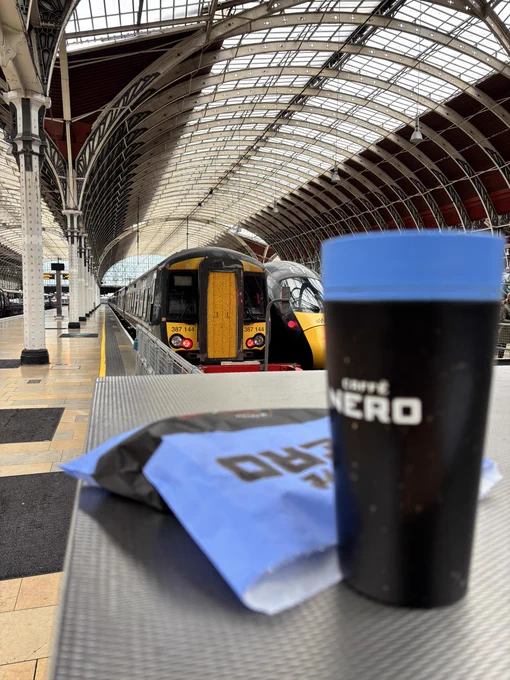 Blue and black coffee cup and blue bag containing a sausage bap on a shelf with a couple of trains in the background - one 387 and one IET