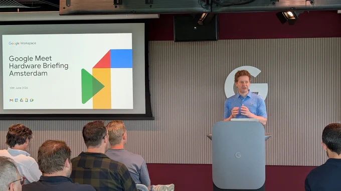 A photo of a man standing at a podium at the front of a room. On the wall behind him, and on the front of the podium, is the letter “G” for “Google”. To his right is a projector screen with the Google Meet logo and the words “Google Meet Hardware Briefing Amsterdam” and the date of the event. In the foreground is a seated audience.