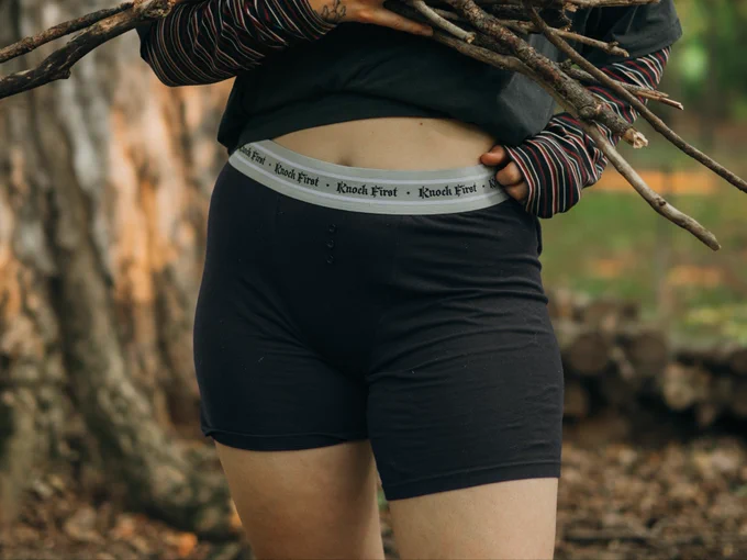Person standing in front of a woodpile wearing black unisex boxers with 3 buttons and “knock first” on the waistband . They are wearing a long sleeve shirt and a baggy black t shirt over it