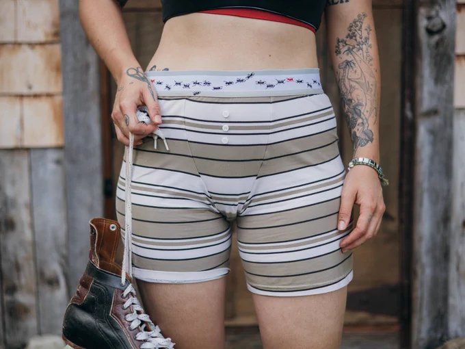 Person standing in front of a wood cabin wearing stripe boxers with 3 buttons and ants on the waistband . They are wearing a red and black bra and holding ice skates
