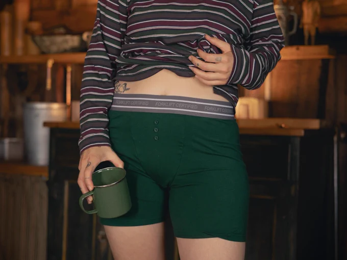 Person standing in front of a cabin kitchen wearing green boxers with 3 buttons and “forklift” on the waistband . They are wearing a striped shirt and holding a green camp mug