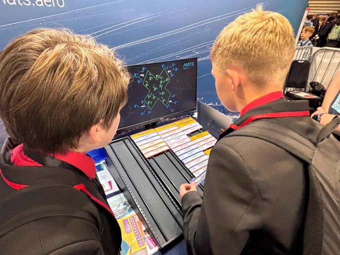 Two people in school uniforms stand at a display, examining a monitor with an air traffic control simulation by NATS. The screen shows various aircraft paths and data.
