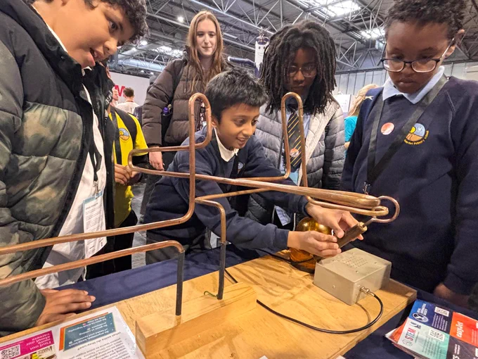 A group of children is engaged with a hands-on exhibit featuring metal wires and a small device on a table. They appear focused and curious, exploring the interactive setup. The setting suggests an educational or science-related event.