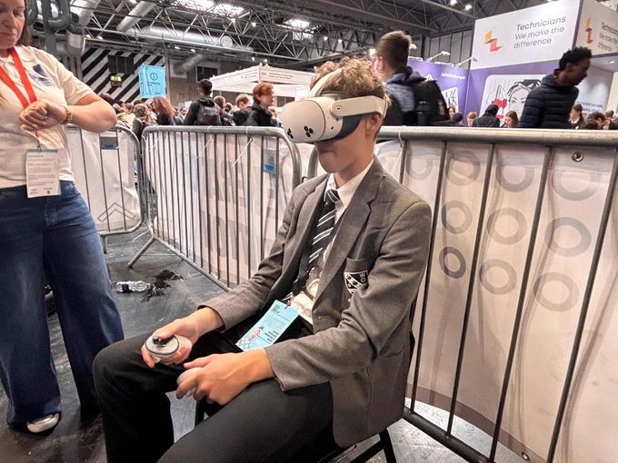 A person seated and wearing a VR headset, holding a controller in each hand. They are dressed in a suit and tie. In the background, there are various booths and banners, including one for "Technicians: We make the difference." The setting appears to be a busy exhibition or conference hall.