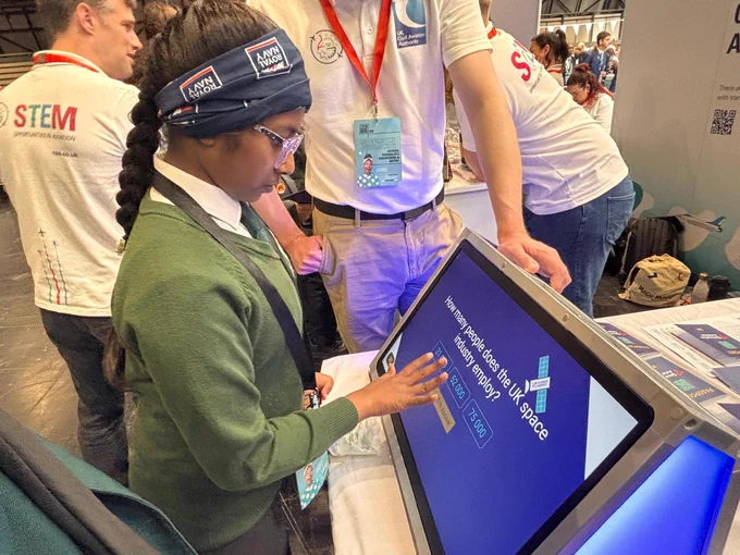 A person in a green sweater interacts with a touchscreen displaying options related to European Space programs. Nearby, someone wearing a white shirt with a red lanyard stands beside them. The event seems related to STEM, with banners and badges featuring "ESA" and "Airbus."