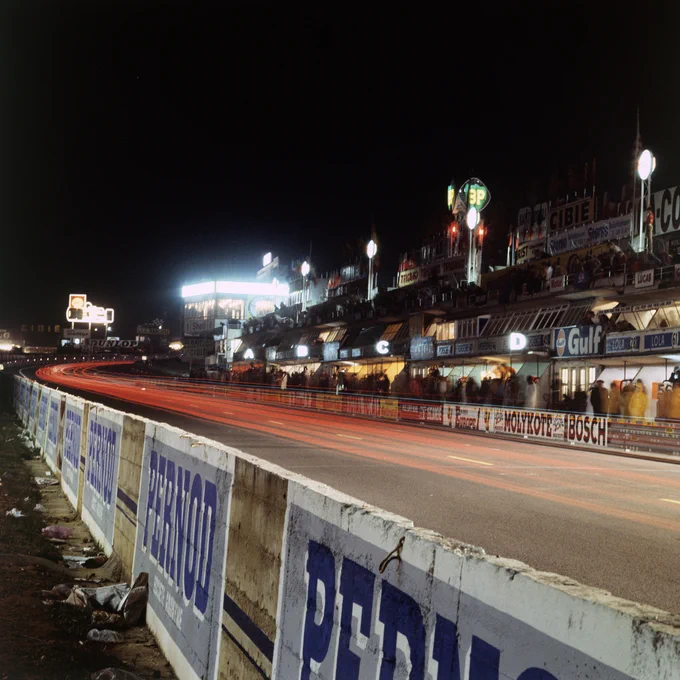 A night shot taken looking along the main straight towards the first corner. Taken with a long exposure, red streaks of light curve rightwards into the corner. Across the track, the pit building glows with various lights, letters indicating garages a bright white, ghostly figures where people have stood and moved. 1973 Le Mans 24 Hours, LAT Images.
