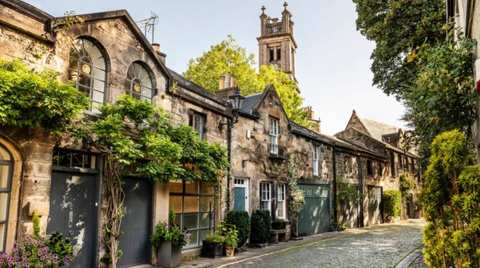 Photo of Circus Lane, in Stockbridge, Edinburgh, There's a row of historic mews properties along a cobbled street, and lots of greenery and a church tower rising behind.