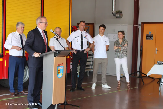 M. le préfet s'exprime derrière un pupitre siglé « SDIS Hautes-Pyrénées » lors d'un événement officiel dans une caserne. Plusieurs personnes, dont le colonel Arnaud Fabre, l'écoutent en arrière-plan à côté d'un camion de pompiers.