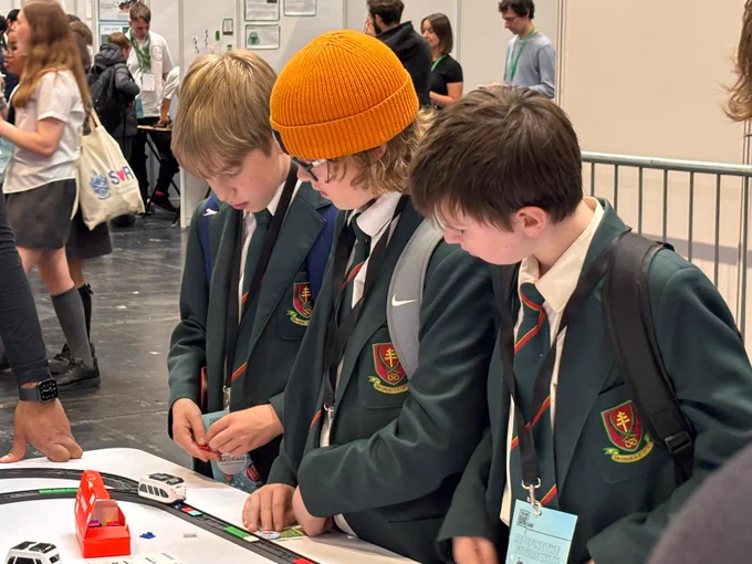 Three students in uniforms with green blazers and red-striped ties are focused on an educational display at a science fair. They're standing behind a table with model cars and a track. Other attendees and a banner with "STEM" can be seen in the background.