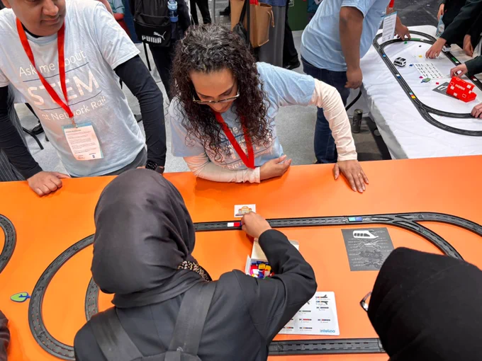 A group of individuals gathered around a bright orange table engaged in a STEM-related activity featuring a small track with miniature vehicles. Several people are wearing lanyards and T-shirts with the word "STEM." A person in the foreground is placing an item on the track. It's at The Big Bang Fair.