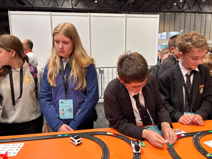 A group of young students is gathered around a small racing track exhibit. They appear engaged with miniature vehicles on the track. The background features a simple booth setup at an event.