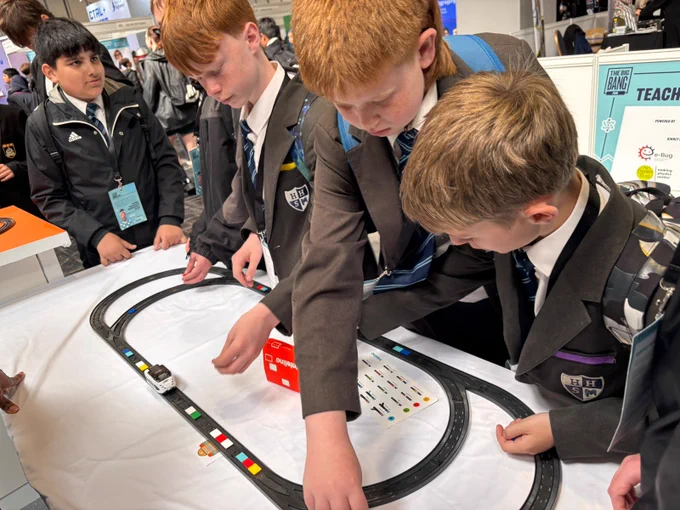 A group of students in school uniforms are gathered around a table with a toy car track. They are observing and engaging with the setup, which includes a sheet displaying various flags and symbols. The scene takes place indoors at an event with other booths and people visible in the background.
