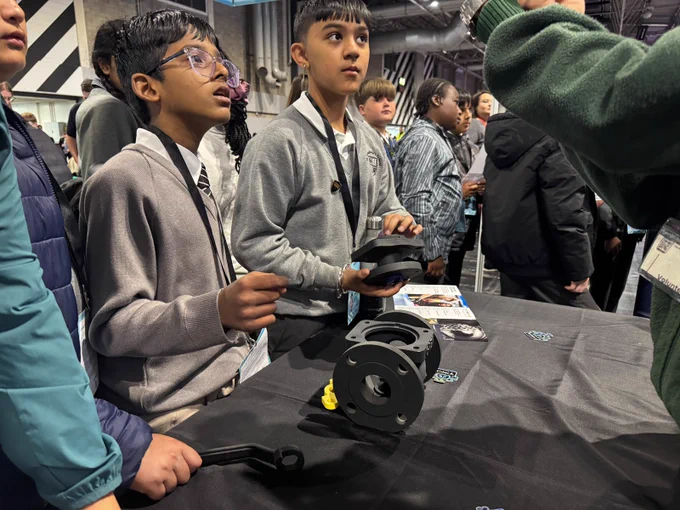 Young students gather around a table. They are attentively observing while one holds a piece of valve, and many wear lanyards. The background shows others engaged in the event.