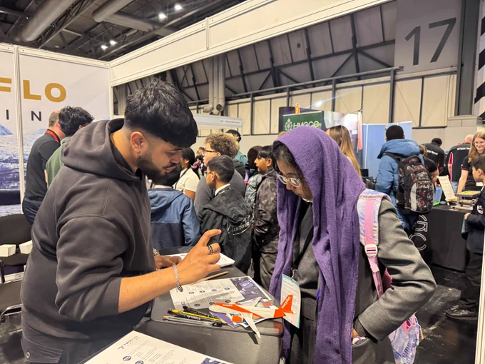 A person is engaged in conversation with another individual at an indoor event. They are looking through displayed materials and a small model airplane. Several people are visible in the background, reading signs and interacting with displays.